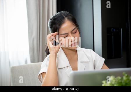 Femme d'affaires asiatique stressée avec les yeux fermés touchant le front assis devant un ordinateur portable au bureau Banque D'Images