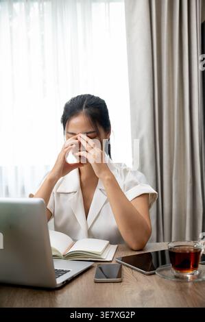 Femme d'affaires asiatique stressée couvrant le visage assis à table avec des gadgets et un cahier dans le bureau Banque D'Images
