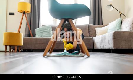 Adolescente hispanique portant des gants en caoutchouc se penchant vers l'avant et utilisant une serpillière dans le nettoyage du plancher en bois sous la chaise dans le salon à la maison Banque D'Images