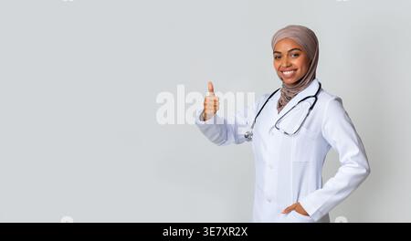 Médecin musulman noir avec stéthoscope portant le hijab montrant les pouces vers le haut tout en se tenant debout sur fond gris dans le studio Banque D'Images