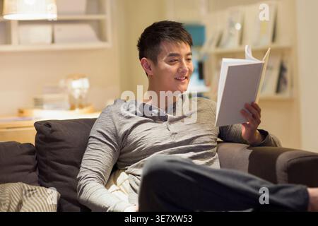 Homme chinois souriant portant le livre de lecture à manches longues grises tout en étant assis sur le canapé à la maison Banque D'Images