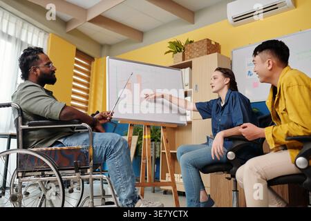 Homme asiatique handicapé en fauteuil roulant pointant bâton sur le tableau blanc discutant chLifestyle avec des collègues assis près de lui dans le bureau pendant la journée Banque D'Images