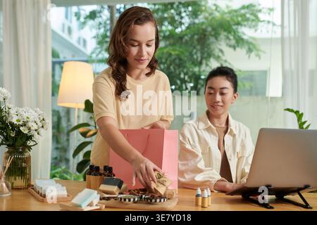 Femme de race mixte mettant cosmétique organique dans un sac en papier avec une amie asiatique travaillant sur ordinateur portable dans le salon à la maison pendant la journée Banque D'Images
