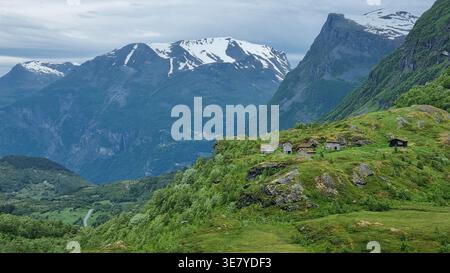 Des sommets enneigés s'élèvent au-dessus de collines verdoyantes et de petites cabanes dans une région reculée de Norvège. Les nuages couvrent le ciel par une journée fraîche. Banque D'Images