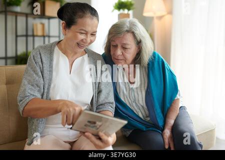 Souriante femme asiatique d'âge moyen montrant un album photo à une amie caucasienne senior assise dans le salon pendant la journée Banque D'Images