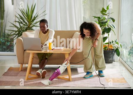 Femme mixte aspirateur tapis tandis que femme noire travaillant sur son ordinateur portable assis sur le canapé dans le salon de l'appartement Banque D'Images