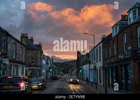 Une scène de rue pittoresque à Innerleithen, en Écosse, baignée dans la lueur chaude d'un coucher de soleil spectaculaire avec des nuages vibrants au-dessus de la tête et des voitures sur la route Banque D'Images