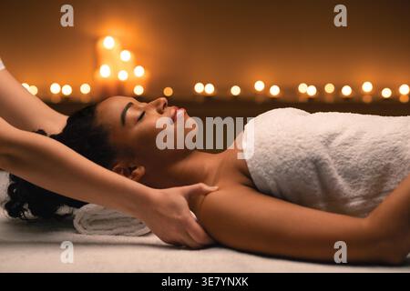 Femme noire avec les yeux fermés envelopper dans une serviette ayant massage de l'épaule couché sur le lit près de bougies allumées dans le spa Banque D'Images