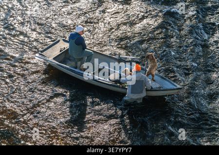 Deux hommes en cuissardes et chapeaux pêchent depuis un petit bateau avec deux chiens, l'un debout et regardant au-dessus de l'eau Banque D'Images