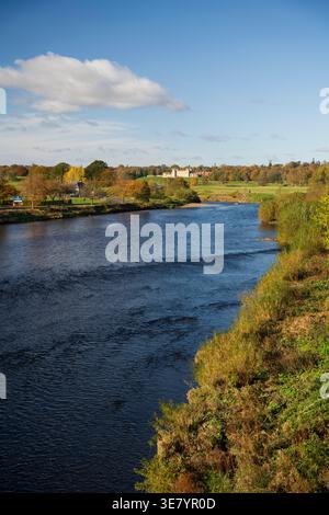 Une large rivière coule à travers un paysage parsemé d'arbres affichant un feuillage d'automne vibrant, menant à un grand domaine sous un ciel bleu avec des nuages Banque D'Images