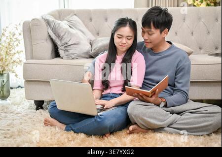 Homme asiatique tenant le livre aidant la petite amie asiatique travaillant sur ordinateur portable assis sur le tapis de sol avec les jambes croisées appuyées contre le canapé dans le salon à ... Banque D'Images