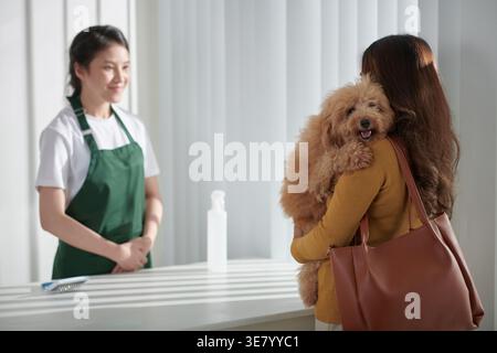 Propriétaire asiatique portant son chien et parlant à la toiletteuse asiatique tout en se tenant debout dans le salon de toilettage Banque D'Images