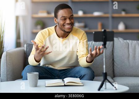 Souriant Black Man faisant du streaming en direct à l'aide d'un smartphone sur trépied donnant leçon en ligne assis sur un canapé près de la table avec tasse à café et livre dans Livin... Banque D'Images