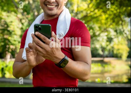 Image recadrée de l'homme thaïlandais dans des vêtements de fitness à l'aide d'un smartphone tout en se tenant debout avec une serviette blanche autour de son cou dans le parc le jour ensoleillé Banque D'Images