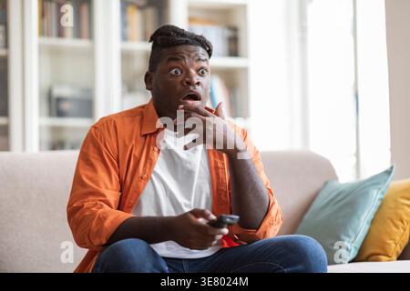 Homme noir choqué touchant le menton tenant la télécommande tout en regardant la télévision assis sur le canapé dans le salon à la maison Banque D'Images