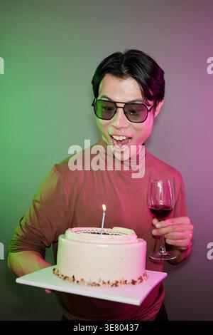Joyeux homme asiatique portant des lunettes de soleil tenant verre de vin et gâteau avec bougie allumée debout contre le mur avec des néons verts et violets dans St... Banque D'Images