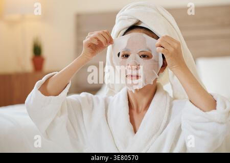 Femme asiatique avec serviette enroulée autour de la tête mettant masque de feuille assis sur le lit après la douche du matin à la maison Banque D'Images