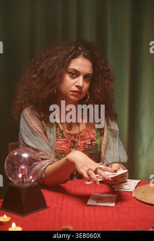 Diseuse de fortune féminine de race mixte étalant des cartes de tarot sur la table tout en étant assise avec une boule de cristal à l'intérieur Banque D'Images