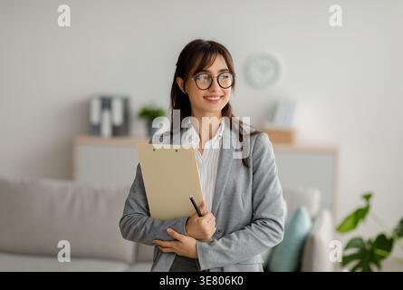 Femme du moyen-Orient portant des lunettes regardant loin tenant une planche à pince tout en se tenant debout dans le salon à la maison Banque D'Images
