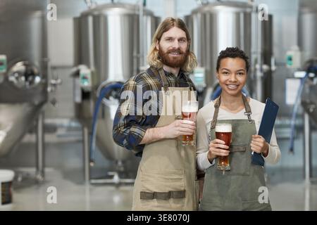 Femmes mixtes et hommes du caucase portant des tabliers tenant un verre de bière tout en se tenant debout dans l'usine de brassage Banque D'Images