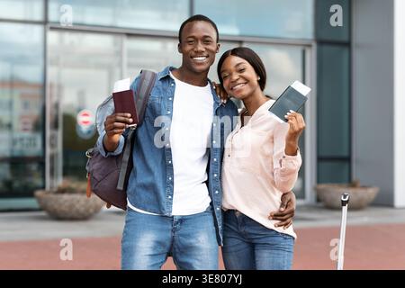 Couple noir souriant avec sac à dos tenant des passeports avec billet d'avion debout près l'un de l'autre à l'extérieur de l'aéroport Banque D'Images