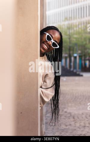 Femme noire avec des tresses portant des lunettes de soleil debout et jetant un coup d'œil hors du pilier du bâtiment dans la ville pendant la journée Banque D'Images