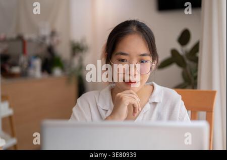 Femme asiatique portant des lunettes touchant le menton à l'aide d'un ordinateur portable assis à la table dans le café Banque D'Images