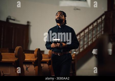 Prêtre noir avec moustache et barbe tenant des perles de chapelet et la Bible regardant vers le haut en priant tout en se tenant debout dans l'allée de l'église Banque D'Images