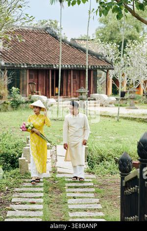 Femme asiatique dans des vêtements traditionnels portant un chapeau tenant des fleurs de lotus tout en marchant avec son petit ami dans le jardin pendant la journée Banque D'Images