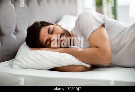 Homme arabe avec la barbe portant une chemise blanche dormant sur le lit dans la chambre à coucher à la maison Banque D'Images