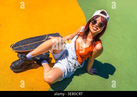 Femme asiatique portant casquette et lunettes de soleil assis avec skateboard sur le sol dans le parc le jour ensoleillé Banque D'Images