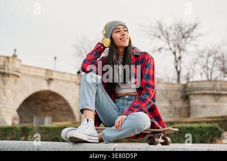 Femme vénézuélienne avec de longs cheveux noirs portant un bonnet écoutant de la musique sur des écouteurs regardant loin tout en étant assise sur le skateboard en ville pendant la journée Banque D'Images