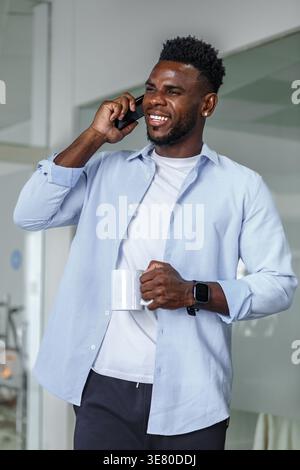 Souriant homme d'affaires noir tenant une tasse de café et ayant un appel téléphonique regardant loin tout en se tenant debout dans le bureau Banque D'Images