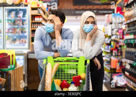 Couple moyen-oriental portant un masque facial penchant leur bras sur le chariot tout en marchant dans le supermarché Banque D'Images