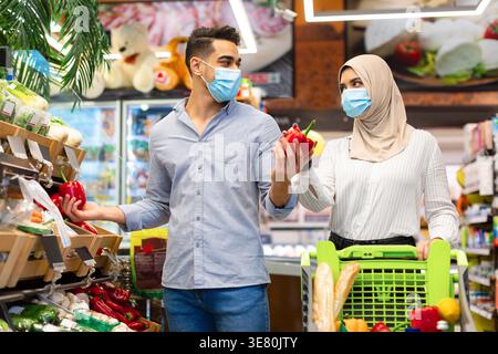 Couple souriant du moyen-Orient portant un masque facial choisissant des légumes sur des étagères tout en se tenant debout avec un chariot dans un supermarché Banque D'Images
