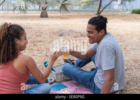 Couple hispanique griller des boissons tout en étant assis sur une couverture de pique-nique au parc Banque D'Images