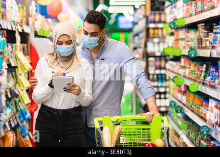 Couple souriant du moyen-Orient portant des masques de visage vérifiant la liste d'épicerie sur le carnet tout en se tenant debout avec le chariot près des étagères de légumes dans le supermarché Banque D'Images