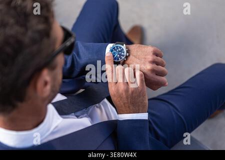 Homme d'affaires hispanique dans des lunettes de soleil vérifiant l'heure tout en étant assis sur le banc sur la rue pendant la journée Banque D'Images