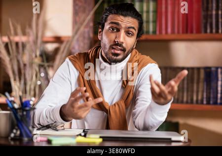 Enseignant indien dans des écouteurs gestuels mains enseignant en ligne assis à table dans la bibliothèque Banque D'Images