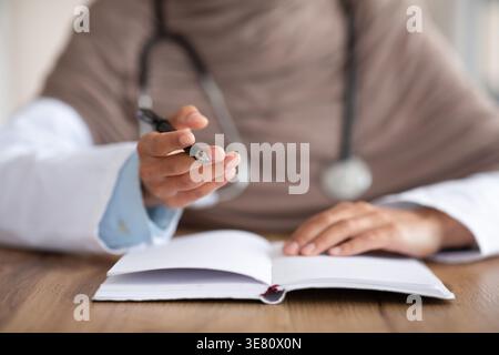 Image recadrée d'une femme médecin du moyen-Orient portant le hijab et le stéthoscope autour de son cou prenant des notes tout en étant assise à table dans la clinique Banque D'Images