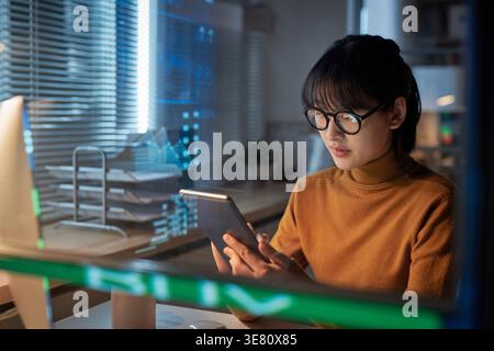 Femme d'affaires asiatique concentrée portant des lunettes en utilisant la tablette assis à table dans le bureau la nuit Banque D'Images