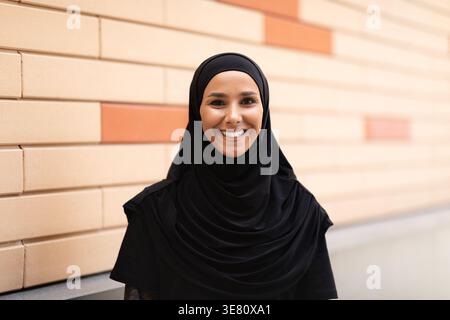 Femme musulmane du moyen-Orient souriante portant un hijab noir debout devant un mur de briques dans la rue pendant la journée Banque D'Images
