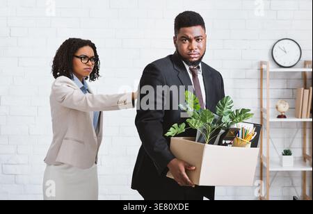 Patron féminin noir virant un employé masculin portant une boîte de choses personnelles dans le bureau Banque D'Images