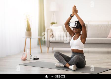 Femme noire avec des vêtements de sport bouclés et casque VR méditant sur tapis dans le salon à la maison Banque D'Images