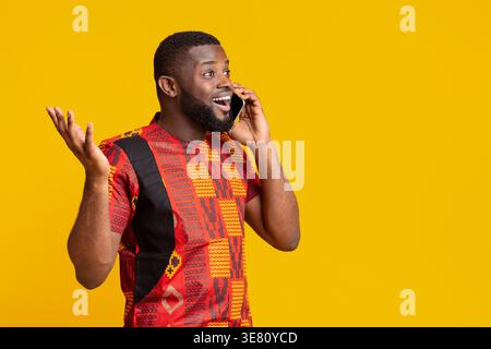 Homme noir surpris regardant loin ayant un appel téléphonique debout avec la main levée sur fond jaune dans le studio Banque D'Images