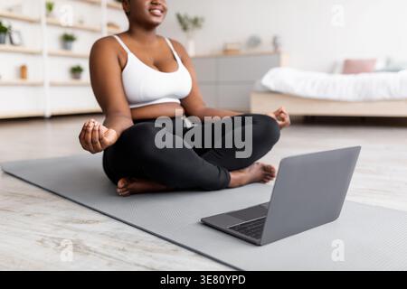 Curvy femme noire assise dans la pose de lotus sur le tapis de yoga près de l'ordinateur portable à la maison Banque D'Images