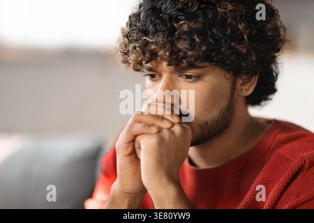 Homme asiatique inquiet avec les mains serrées près de sa bouche regardant loin assis sur le canapé à la maison Banque D'Images