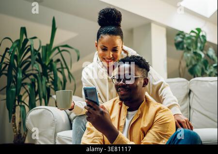 Homme noir assis sur le canapé partageant le smartphone avec femme mixte debout derrière à la maison Banque D'Images