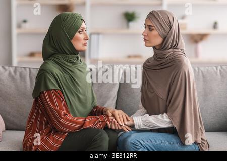 Mère musulmane moyen-orientale d'âge moyen et fille en hijab se tenant les mains se soutenant tout en étant assis sur le canapé dans le salon à la maison Banque D'Images