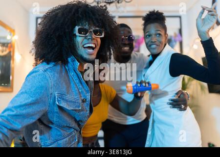 Homme hispanique avec afro portant des jeans en denim et des lunettes de soleil s'amuser à chanter karaoké et danser avec des amis noirs masculins et féminins pendant la fête de la maison Banque D'Images